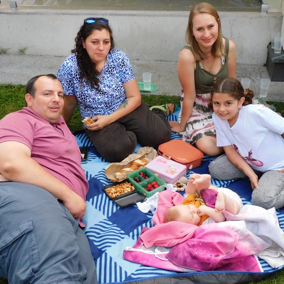 Eine Familie genießt ein Picknick auf einer Rasenfläche. Ein Baby schläft auf einer Decke, während die anderen essen und lächeln.