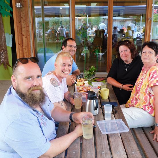 Fünf Menschen sitzen lächelnd um einen Holztisch im Freien. Ein Mann mit Bart und Sonnenbrille auf dem Kopf hält ein Glas. Eine Karaffe und Tassen stehen auf dem Tisch. Eine Frau in einem Blumenshirt sitzt neben einer Frau in einem schwarzen Shirt. Hinter ihnen sind Glastüren, die auf eine Terrasse führen.