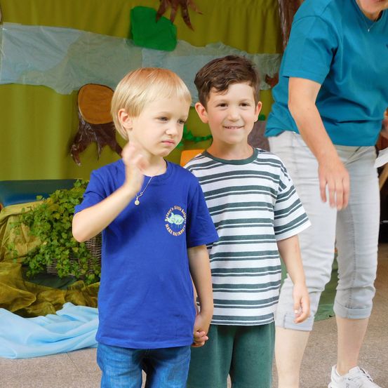 Zwei junge Jungen stehen eng beieinander, einer in einem blauen T-Shirt und der andere in einem gestreiften T-Shirt, mit einer Frau in einem blauen T-Shirt und weißen Hosen hinter ihnen.