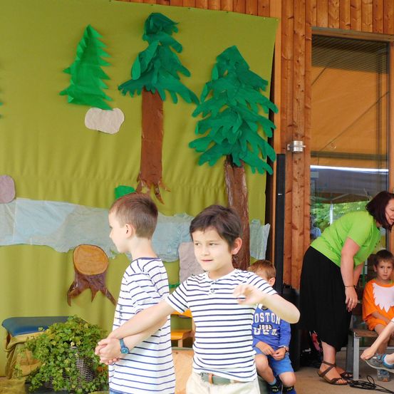 Zwei junge Jungen stehen mit erhobenen Händen vor einer Wand mit Papierbäumen. Eine Frau in Grün und ein Kind in Orange sitzen in der Nähe.