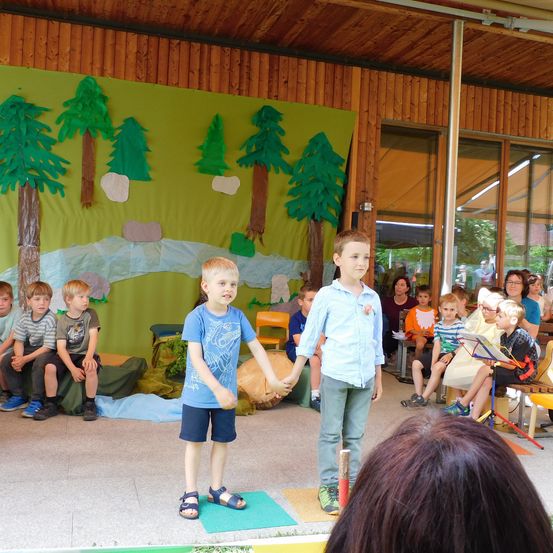 Zwei Jungen stehen Hand in Hand auf der Bühne. Dahinter zeigt eine Kulisse eine Waldszene. Zuschauer, darunter Kinder und Erwachsene, sitzen auf Stühlen, einige beobachten aufmerksam.