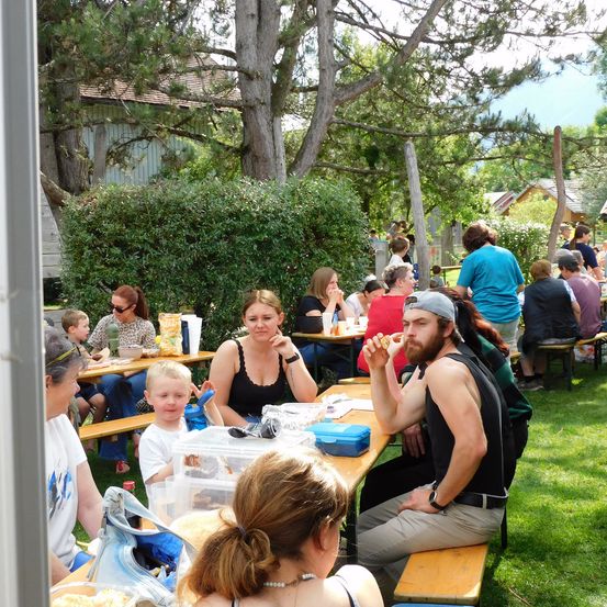 Eine Gruppe von Menschen sitzt auf Bänken in einem Park. Einige essen, andere reden. Ein Junge schaut etwas an.