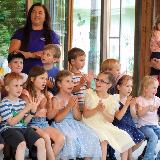 Eine Gruppe von Kindern sitzt zusammen, klatscht und lächelt. Eine Frau in einem blauen T-Shirt steht hinter ihnen und leitet die Aktivität. Die Kinder tragen verschiedene Kleidungsstücke, und ein Baum ist durch ein Fenster sichtbar.
