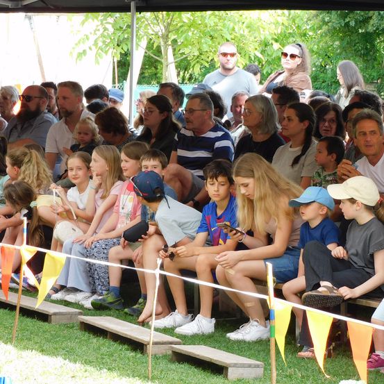 Eine Menschenmenge, bestehend aus Erwachsenen und Kindern, sitzt in Reihen in einem Außenbereich. Einige von ihnen schauen auf ihre Telefone. Bunte Fahnen befinden sich am Zaun. Bäume und Pflanzen sind im Hintergrund.
