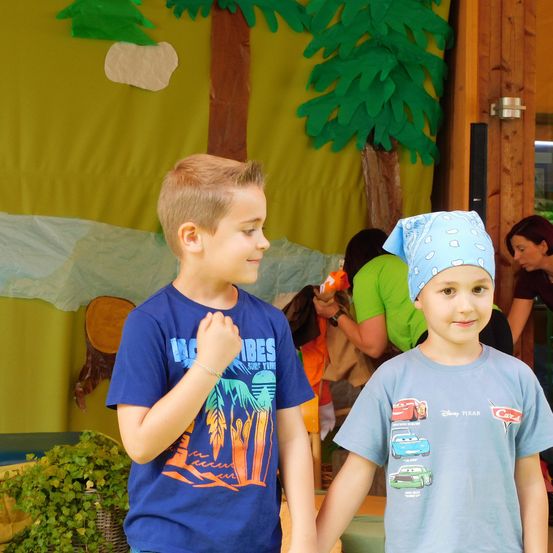 Zwei junge Jungen stehen in einem Raum mit grünen Dekorationen und Bäumen. Ein Junge trägt ein blaues T-Shirt mit einem gedruckten Design und einem Bandana. Der andere Junge hat ein blaues T-Shirt mit Autos bedruckt. Im Hintergrund sind Menschen und ein Baumstamm zu sehen.