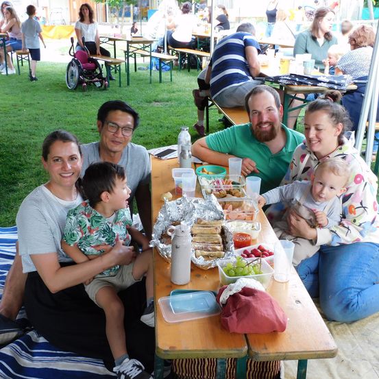 Eine Familie sitzt auf einer Decke auf dem Rasen und macht ein Picknick. Sie haben Essen auf dem Tisch, einschließlich Sandwiches, Obst und Getränke. Eine andere Familie sitzt im Hintergrund auf einer Bank.