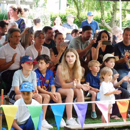 Eine Gruppe von Menschen auf Tribünen mit Wimpelketten. Einige klatschen und andere benutzen ihre Telefone. Ein Mädchen mit langen blonden Haaren ist vorne.