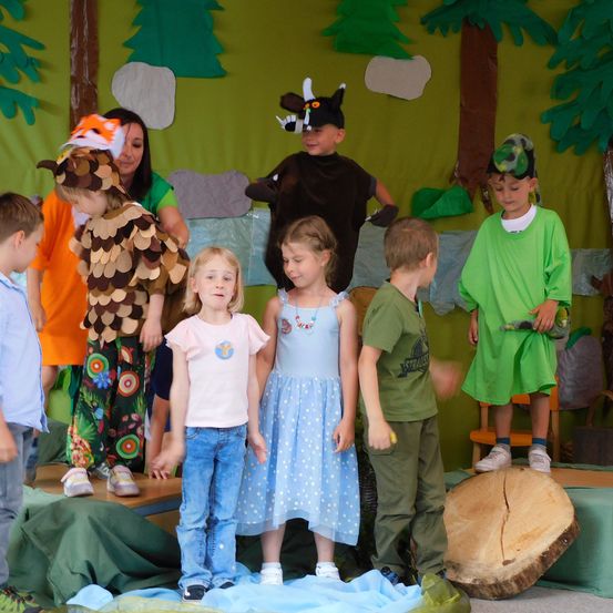 Eine Gruppe von Kindern in verschiedenen Kostümen steht auf einer Bühne in einer waldartigen Umgebung. Einige tragen Tierkostüme, andere grün. Ein Baumstamm liegt auf dem Boden.