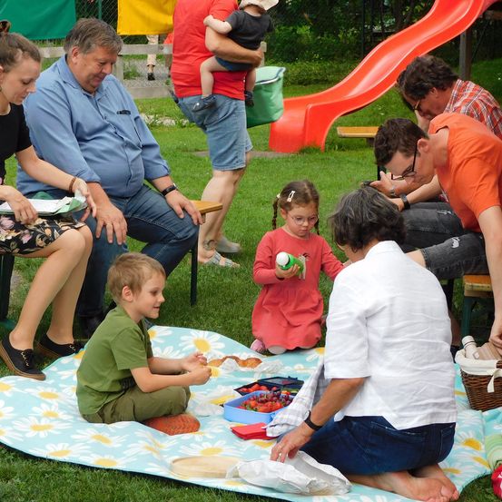 Eine Gruppe von Menschen, darunter Kinder, sitzt auf einer Picknickdecke auf einem Grasfeld. Einige essen, während andere Essen halten. Ein Rutsch ist im Hintergrund.