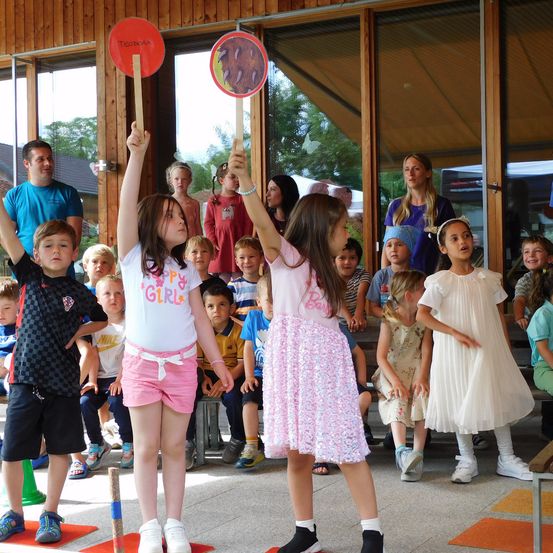 Zwei junge Mädchen halten Schilder als Teil einer Gruppenaktivität in einem Saal. Andere Kinder und Erwachsene beobachten. Der Saal hat Holzwände und Glastüren.