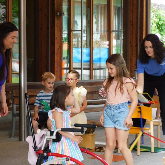 Eine Gruppe von Kindern und Erwachsenen ist draußen. Die Kinder spielen und interagieren miteinander. Ein Mädchen steht neben einem roten Fahrrad.