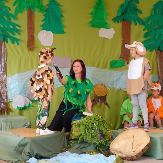 Eine Frau hält ein Mikrofon und spricht zu zwei Kindern in Wald-Kostümen. Dahinter ist eine Kulisse aus Papierbäumen und einem Bach zu sehen. Andere Kinder sitzen auf einer Bank, ebenfalls in Kostümen.