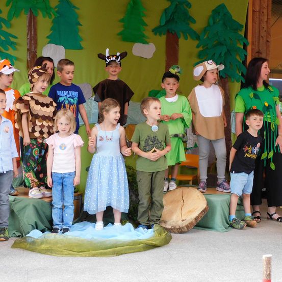 Kinder in verschiedenen Kostümen stehen auf einer Bühne mit einem Waldhintergrund. Einige tragen Tierkostüme, während andere grüne Blätter tragen. Ein Mädchen hält ein blaues Tuch und ein Baumstamm ist vor ihnen platziert.
