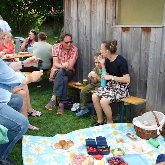 Eine Familie und Freunde genießen ein Picknick an einem sonnigen Tag. Sie sitzen auf Bänken und einer Picknickdecke, essen Essen und Getränke. Eine Holzwand ist im Hintergrund.