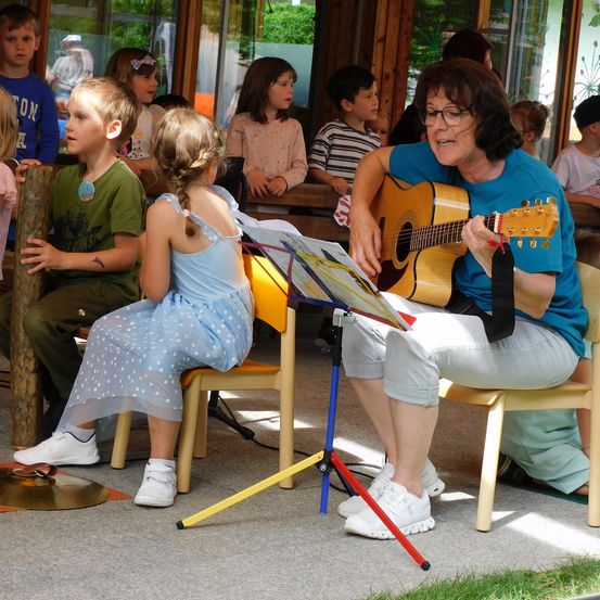 Eine ältere Frau spielt Gitarre vor einer Gruppe von Kindern, die auf Stühlen sitzen und zusehen. Die Frau trägt eine Brille und ein blaues T-Shirt. Hinter ihr sitzen Kinder auf einer Bank und schauen zu.