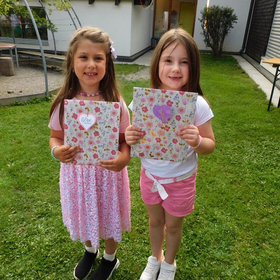 Zwei kleine Mädchen mit blumigen Notizbüchern lächeln für die Kamera auf einem Rasen. Dahinter steht ein Gebäude mit einer grünen Tür.