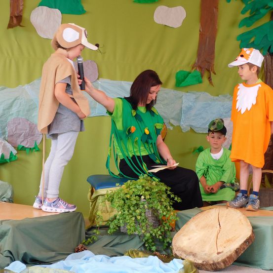 Eine Frau mit einem Mikrofon steht auf einer Bühne mit drei Kindern, eines hält ein Mikrofon, ein anderes liest und das dritte sitzt. Sie tragen Kostüme vor einem grünen Hintergrund.