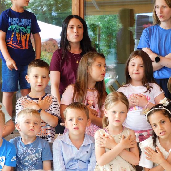 Eine Gruppe von Kindern und zwei Frauen stehen zusammen, wahrscheinlich bei einer Schulveranstaltung. Die Kinder haben ihre Hände vor sich verschränkt. Ein Kind trägt eine Kopfband.