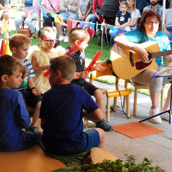 Eine Frau mit Brille spielt Gitarre, während Kinder vor ihr sitzen und zusehen. Ein Kind hält ein rotes Musikinstrument. Sie befinden sich in einem Außenbereich.