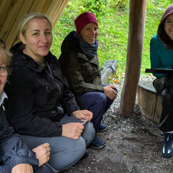 Vier Frauen sitzen im Freien in einem Holzunterstand, lächeln und unterhalten sich. Eine hält ein Tablet, eine andere trägt eine rote Mütze.