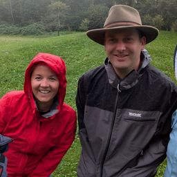 Ein Mann und eine Frau stehen auf einer Wiese und lächeln für ein Foto. Die Frau trägt eine rote Jacke mit Kapuze, während der Mann eine schwarze und graue Jacke trägt. Dahinter befinden sich Bäume und Büsche.