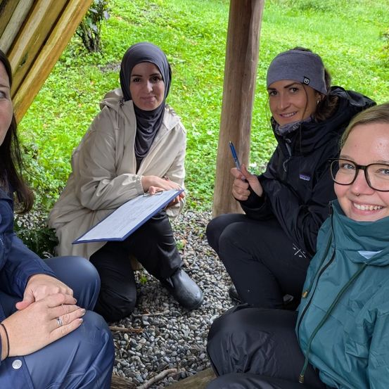 Vier Frauen sitzen lächelnd und interagieren auf Kies vor einer Holzstruktur. Eine hält eine Clipboard, eine andere schreibt mit einem Stift. Sie sind für Outdoor-Aktivitäten gekleidet.