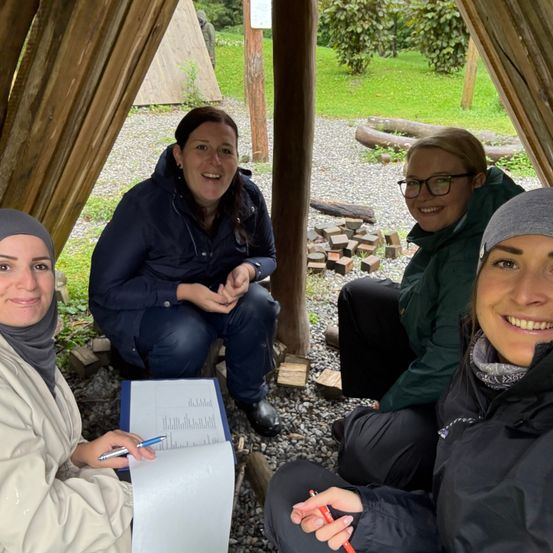 Vier Frauen sitzen in einem Holzunterstand und lächeln für ein Foto. Eine hält eine Mappe und einen Stift. Die Umgebung ist im Freien mit Kies und Pflanzen.