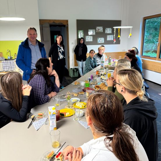 Eine Gruppe von Menschen sitzt um einen langen Tisch in einem Essbereich herum, isst und unterhält sich. Ein Mann in einer blauen Jacke steht lächelnd am Kopf des Tisches. Verschiedene Lebensmittel, darunter Brot, Obst und Getränke, stehen auf dem Tisch. Einige Leute stehen in der Nähe der Tür.