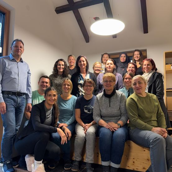 Eine Gruppe von Menschen, darunter Männer und Frauen, lächelt und posiert für ein Foto in einem Raum. Sie sitzen auf einer Holzbank. Der Raum hat weiße Wände und Holzbalken an der Decke. Rechts im Raum steht ein Regal.