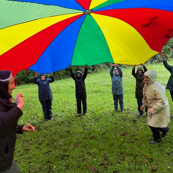 Eine Gruppe von Menschen in Regenmänteln hält ein großes mehrfarbiges Fallschirm im Freien auf einer Wiese.