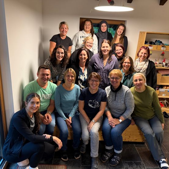 Eine Gruppe von Frauen und Männern posiert für ein Foto in einem Raum, einige sitzen auf einer Bank, andere stehen. Sie alle lächeln.