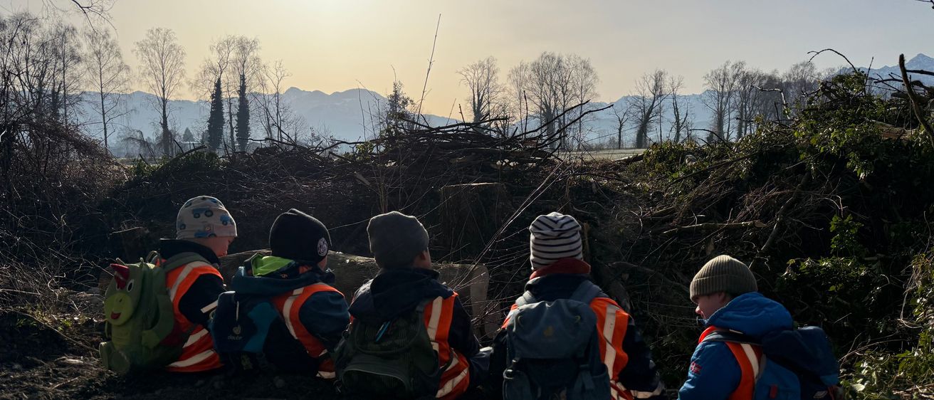 Sechs Kinder in orangefarbenen Westen sitzen auf dem Boden in einem Waldgebiet mit Bergen im Hintergrund.