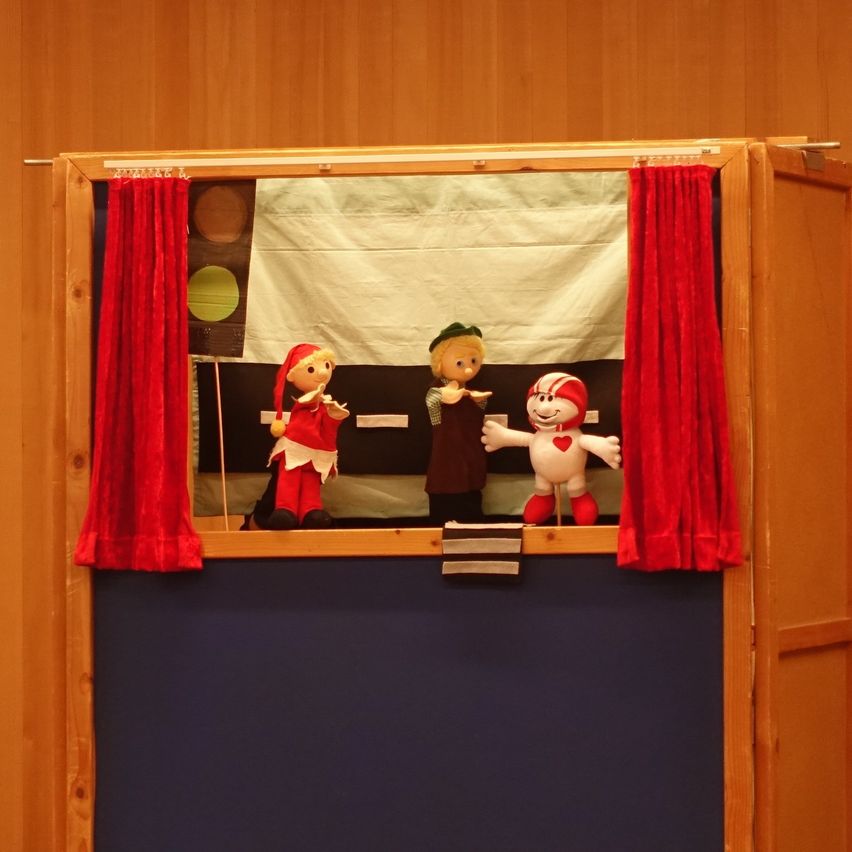 Drei Puppen auf einer hölzernen Bühne mit roten Vorhängen und einem weißen Hintergrund, wahrscheinlich für ein Puppenspiel.