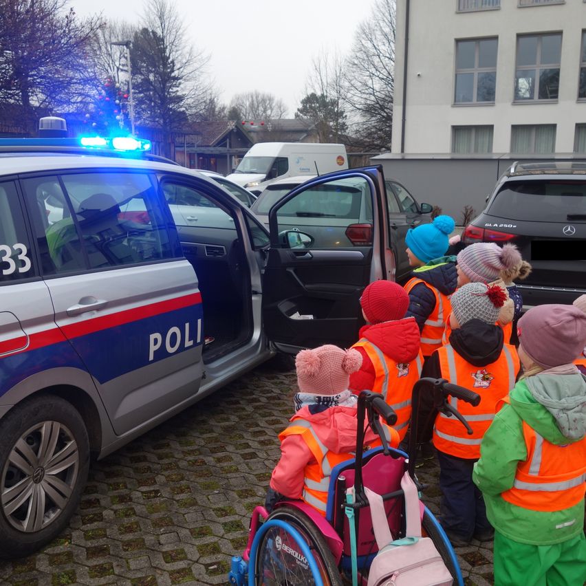 Ein Polizist spricht mit einer Gruppe von Kindern, die orange Westen tragen, einige im Rollstuhl, vor einem Polizeiauto. Bäume und Gebäude sind im Hintergrund zu sehen.