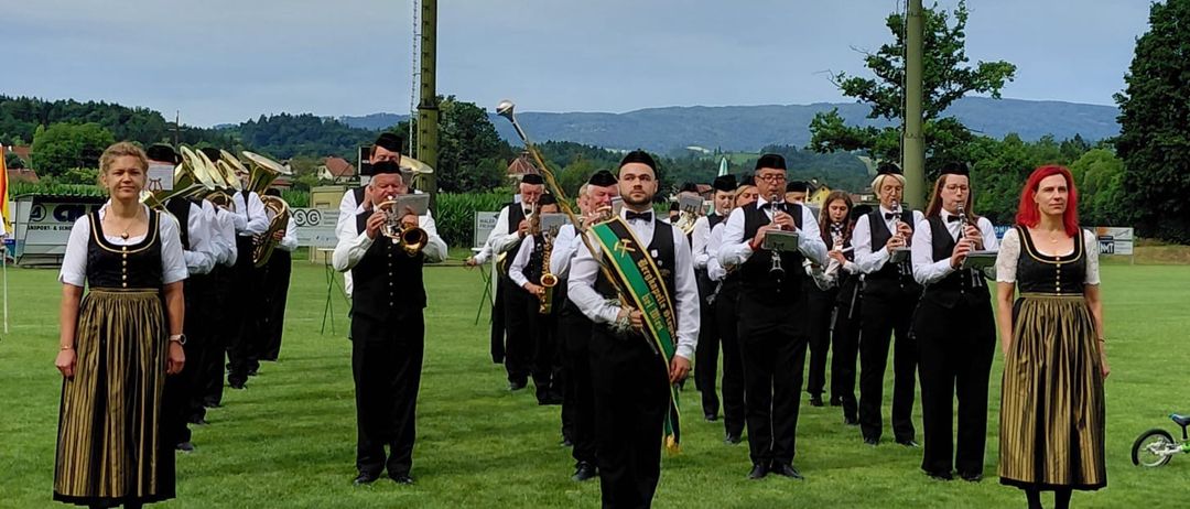 Eine Blaskapelle in traditioneller Kleidung steht auf einem grünen Feld mit Bergen im Hintergrund unter einem bewölkten Himmel.