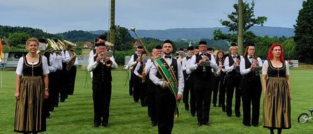Eine Blaskapelle in traditioneller Kleidung steht auf einem grünen Feld mit Bergen im Hintergrund unter einem bewölkten Himmel.