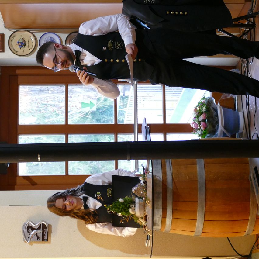 Ein Mann spricht in ein Mikrofon, während er hinter einem Holzfass steht, und eine Frau mit Blumen sitzt darunter. Die Umgebung besteht aus einer Holztür und Fenstern mit Blick auf Bäume draußen.