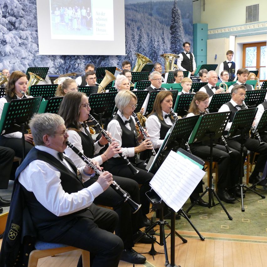 Ein Orchester spielt drinnen. Die Musiker spielen Instrumente wie Trompeten, Posaunen und Klarinetten. Sie sitzen vor Notenständern. Eine Projektionsleinwand zeigt ein Bild von Menschen.