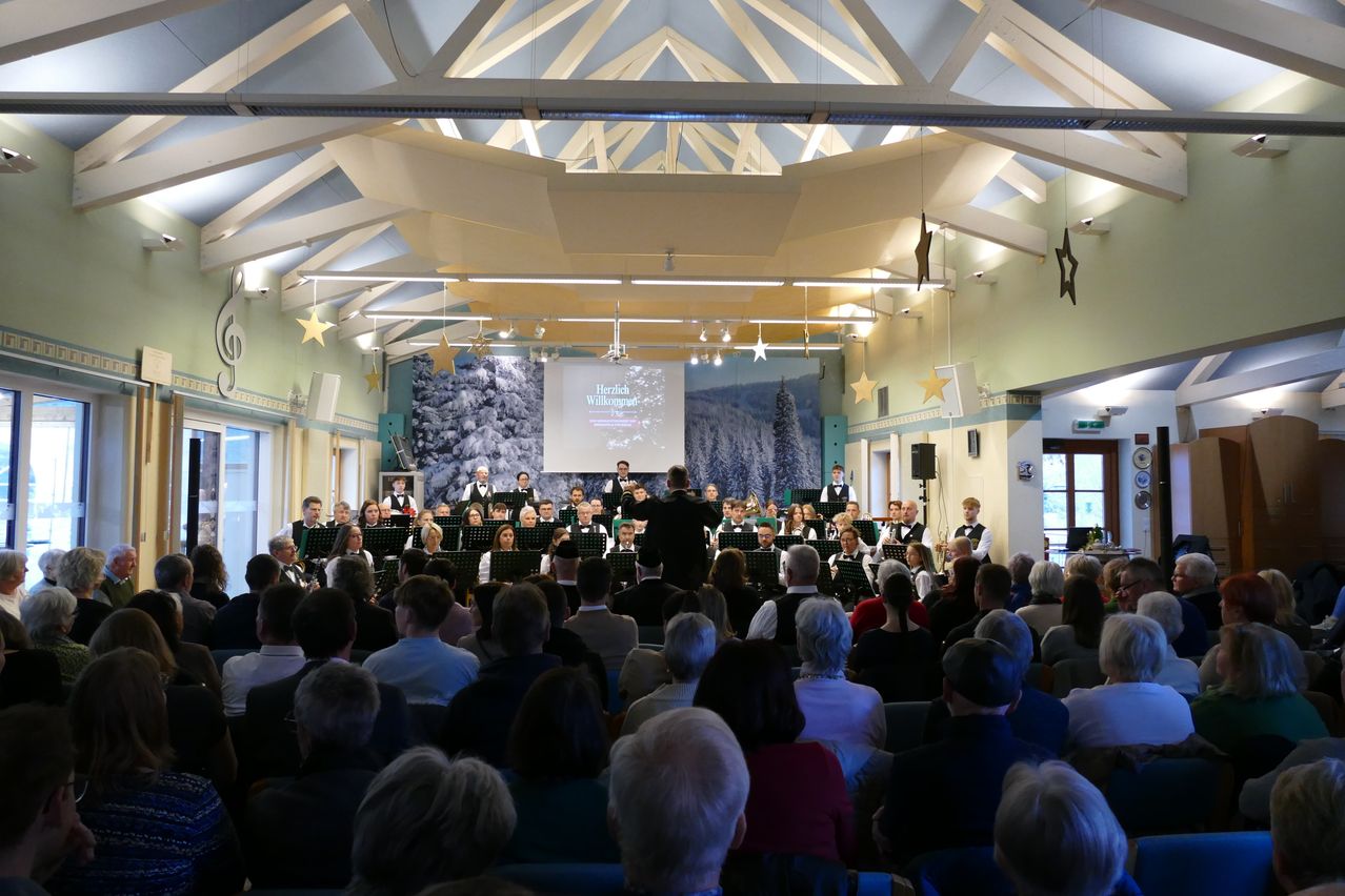 Ein großer Saal mit hohen Decken und einer Projektionsleinwand zeigt eine Musikkapelle, die vor einem Publikum auftritt. Die Kapelle besteht aus Blech-, Holz- und Streichinstrumenten. Das Publikum sitzt in Reihen und beobachtet die Aufführung.