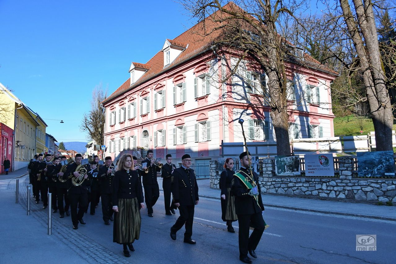 A marching band is performing in front of a pink building with a tree on the side. The band members are dressed in formal attire and playing musical instruments. The building has many windows and a fence on the side.