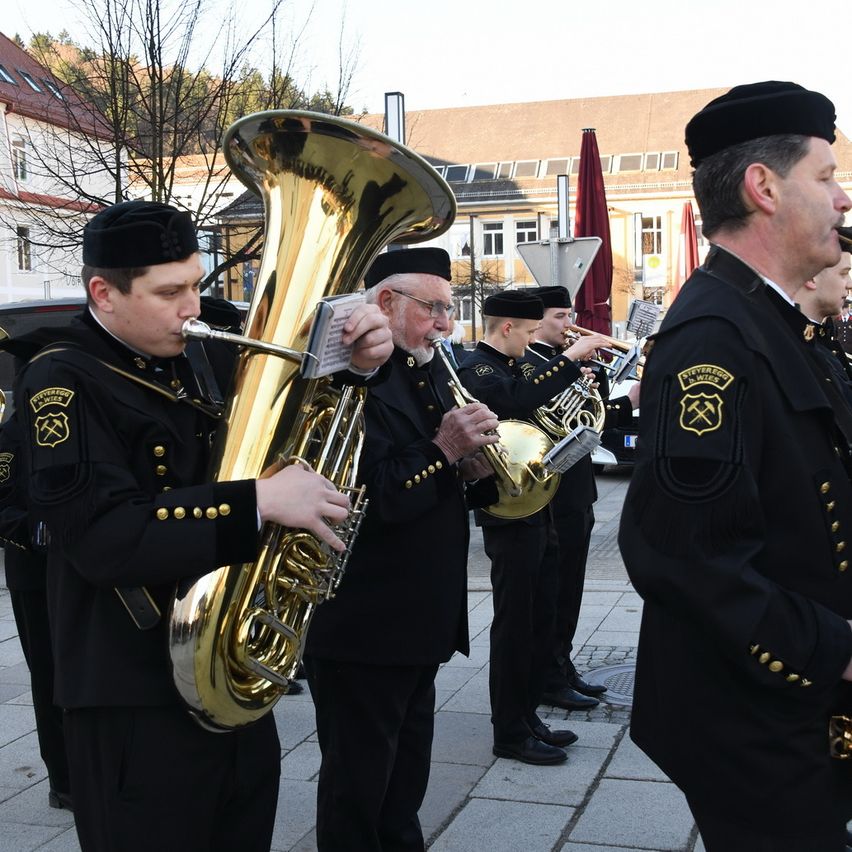 A band of musicians dressed in black uniforms plays instruments on a street, with a man watching them.