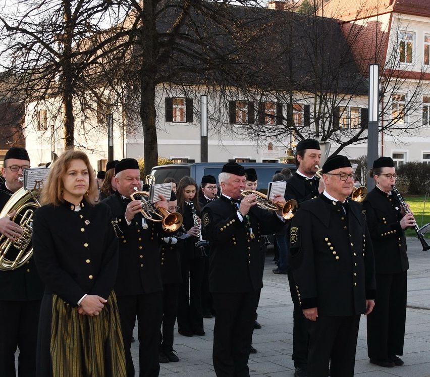 A group of people in uniform, including men and women, are performing with brass instruments in front of a building.