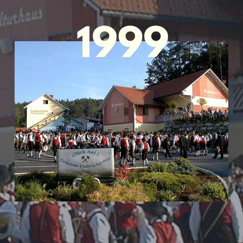 Eine Gruppe von Menschen in traditionellen Kostümen zieht in einem Umzug vor Gebäuden mit der Überlagerung '1999'.