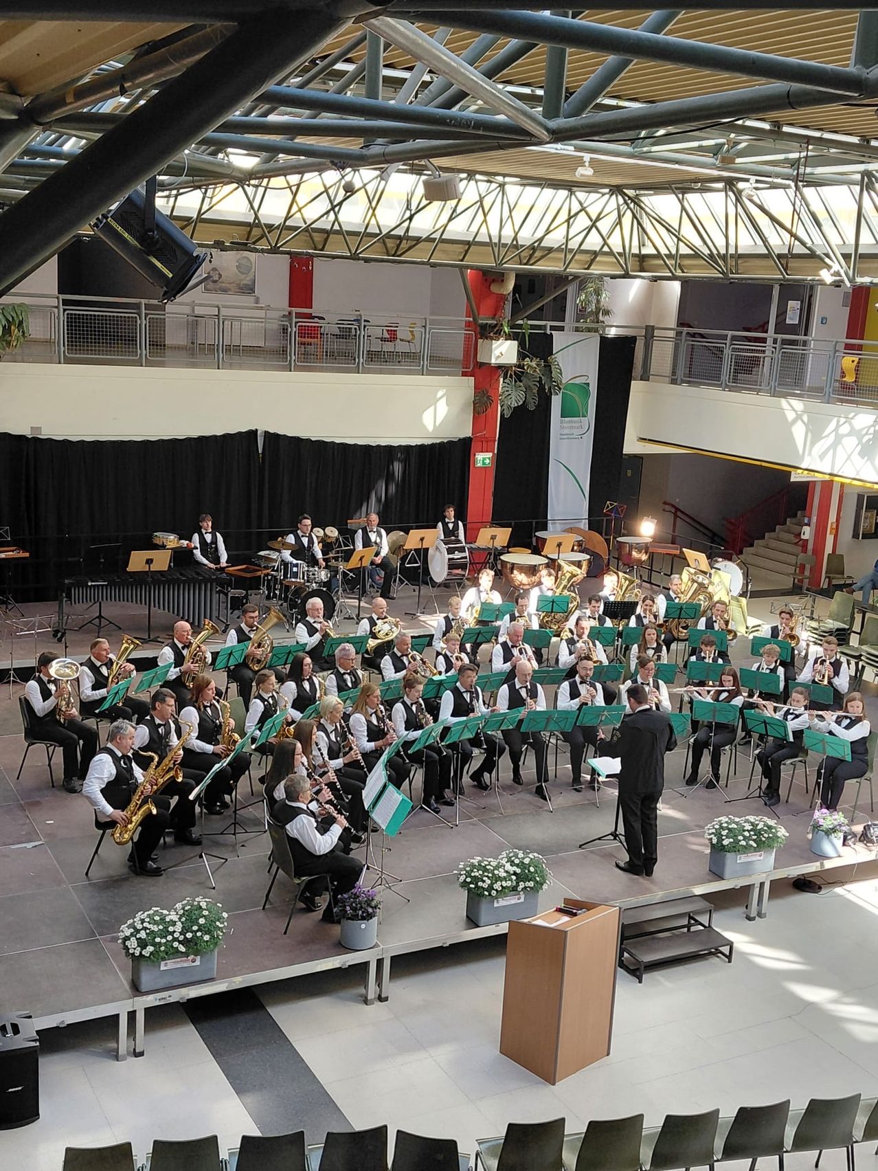 Ein großes Orchester spielt auf der Bühne mit verschiedenen Instrumenten. Die Bühne ist mit weißen Blumen in Töpfen geschmückt. Das Publikum sitzt im Hintergrund.