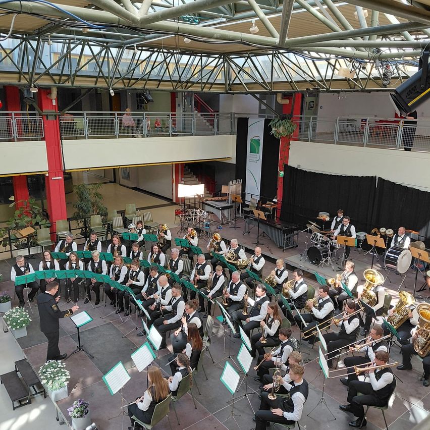 Ein Orchester spielt auf einer Bühne mit einem Dirigenten, verschiedenen Instrumenten wie Posaunen und sitzenden Musikern. Die Bühne ist von einem Metallgeländer eingefasst, und das Publikum sitzt auf Balkonen.