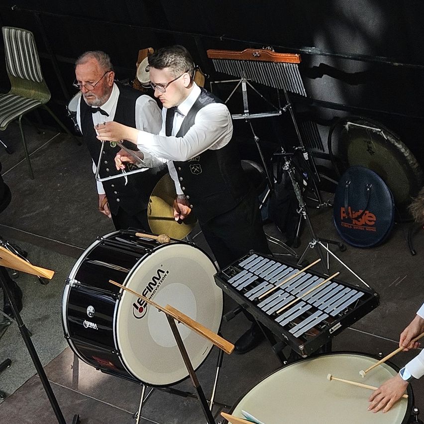 Zwei Männer sind in einem Musikzimmer, einer hält ein Dreieck und der andere einen Drumstick. Sie spielen eine Trommel und ein Xylophon.