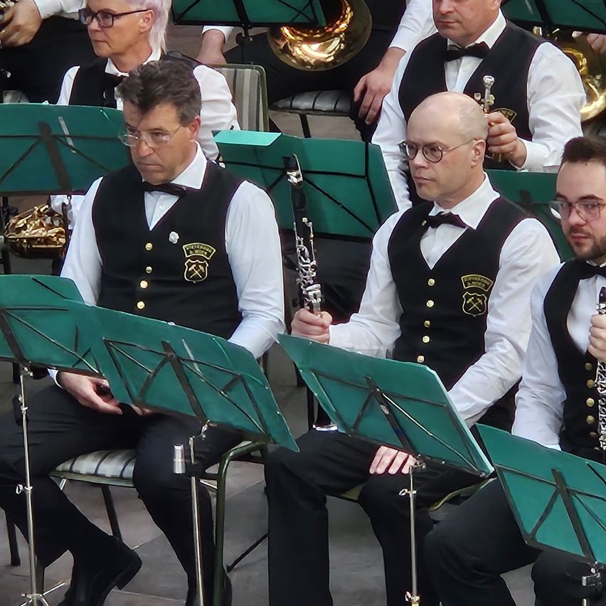 Eine Gruppe von Musikern in formeller Kleidung sitzt vor grünen Notenständern. Ein Mann spielt Klarinette, während ein anderer eine Trompete hält.