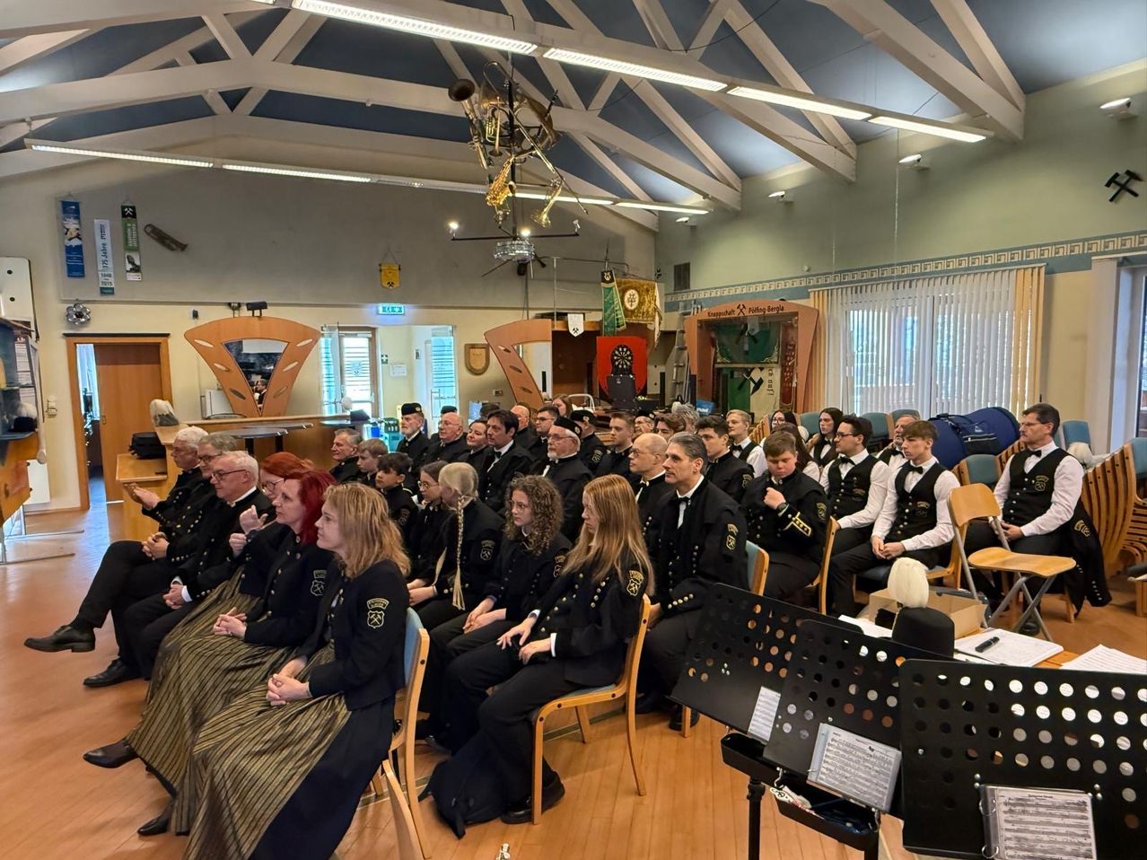 Eine große Gruppe uniformierter Personen sitzt in einem Raum mit Musikinstrumenten und einem Dirigentenpult. Der Raum hat Holzböden und weiße Decken mit hängenden Lampen.