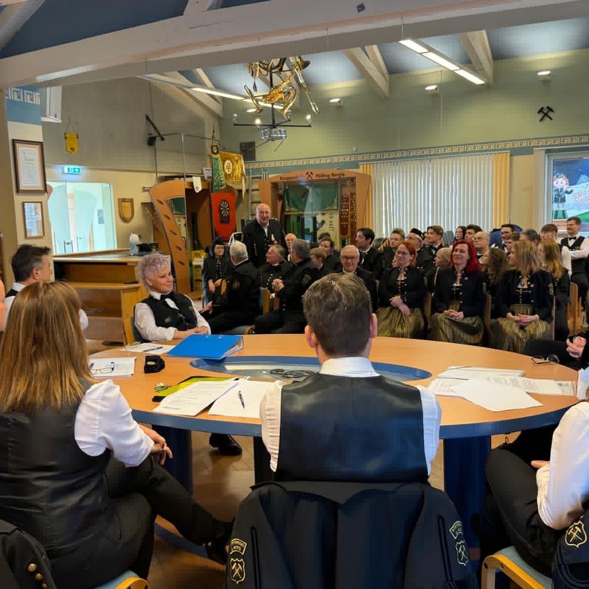 Eine Gruppe von Menschen in Uniformen versammelt sich in einem Saal, wahrscheinlich für eine Besprechung. Sie sitzen um einen Tisch mit Papieren und Stiften herum, und ein Mann in einem weißen Hemd steht vor ihnen. Der Raum hat eine Holzdecke mit Lichtern und ein großes Fenster mit Blick nach draußen.