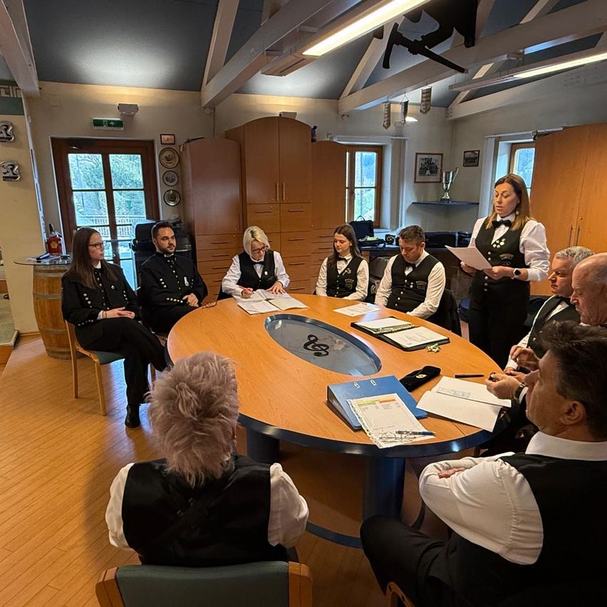 Eine Gruppe von Menschen, die schwarze Westen und weiße Hemden tragen, versammelt sich in einem Raum. Sie sitzen um einen großen ovalen Tisch mit Papieren und Stiften. Die Frau in der Mitte hält ein Blatt Papier.